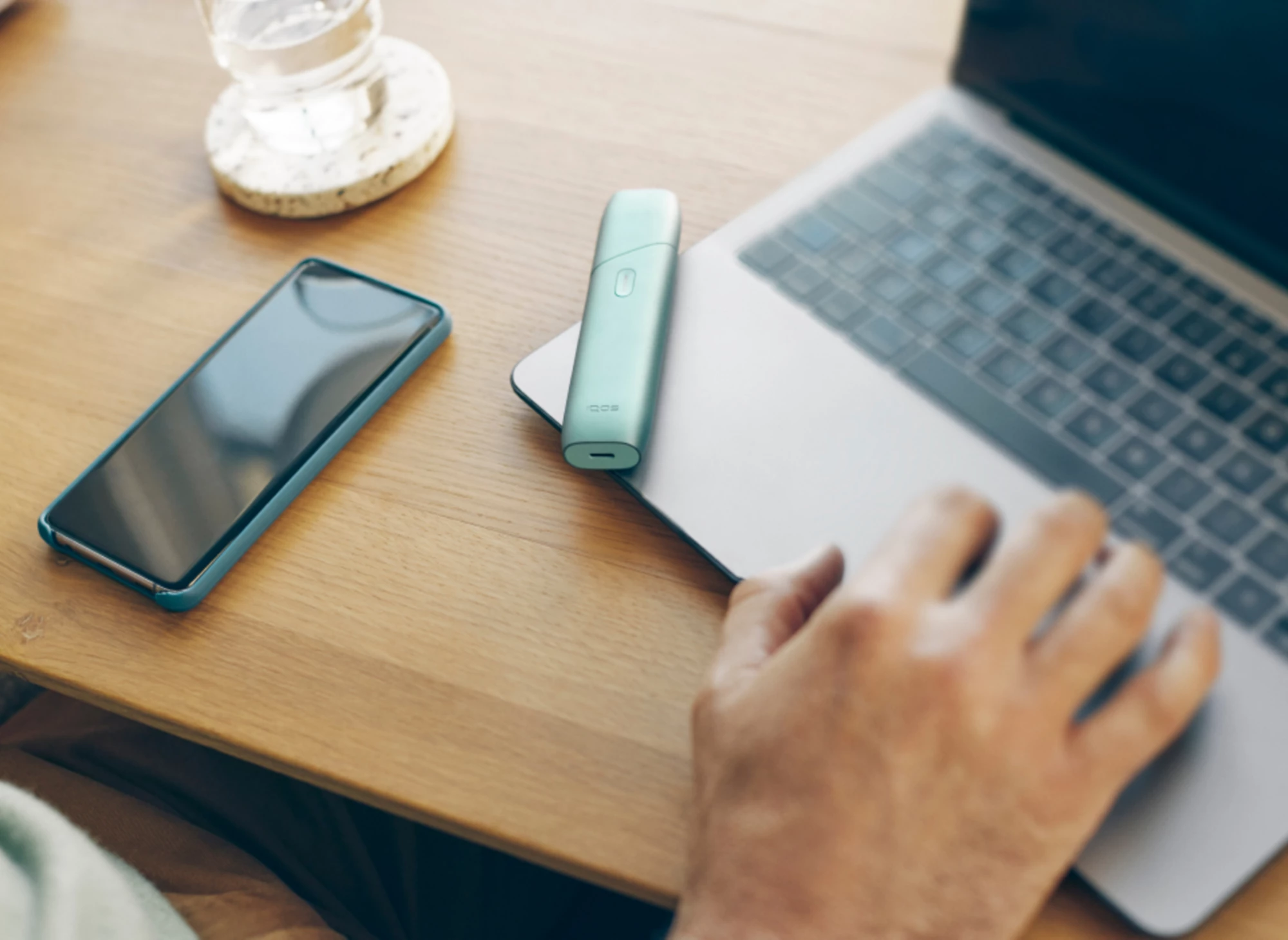 Zariadenie IQOS ILUMA ONE umiestnené na notebooku vedľa mobilného telefónu na stole.